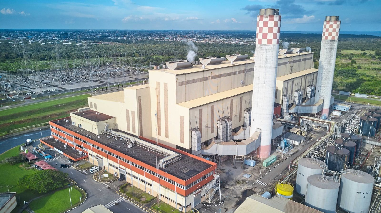 Aerial view of a power station with two tall red-and-white checkered chimneys, a large turbine hall, fuel and water storage tanks, and a switchyard beside it.
